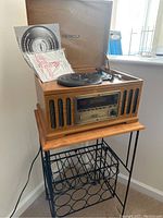 Detrola record player with open lid, displayed on metal stand with wooden top and record storage rings below, including instruction manuals inside lid.
