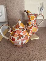 Teapot and pitcher side by side showing autumn leaves decorative pattern and gold trim details.