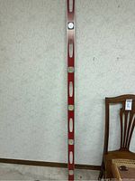 Full length image of a red aluminum level with four bubble vials and oval cutouts along its length, standing against wallpaper.