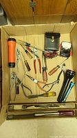 Photo of assorted pliers, screwdrivers, flashlights and an electrical tester all placed inside a cardboard box.