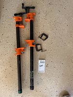 Two large bar clamps with orange adjustment handles and two small black metal C-clamps on speckled floor.