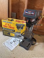 Full side view of drill press with original box and manual on the floor.