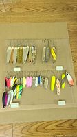 Photo showing an assortment of about 29 fishing lures laid out on a flat surface, including metal spoons in silver, gold, and painted finishes, and colorful fishing spoons and blades with treble hooks.