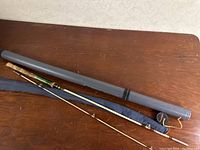 Fishing pole disassembled showing three-piece bamboo rod with green handle and blue fabric sleeve next to rigid grey fishing rod case.