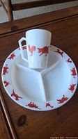 White divided plate with three compartments, decorated with red silhouettes of farm animals such as pigs and roosters around the rim, and matching child's cup with red animal designs.