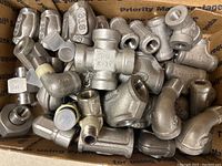 Box filled with various metal plumbing fittings in silver/gray, including elbows, tees, and couplings.