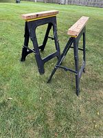 Two black plastic folding sawhorses with wooden tops, set up on grass outdoors, shown from angled side view.