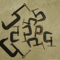 Photo showing 11 assorted small metal C-clamps arranged on a beige surface, various sizes, mostly black coated metal with silver screws, one brass colored clamp