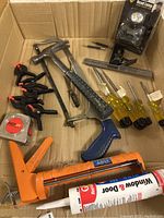 Wide view of the lot including caulking gun, screwdrivers, clamps, headlamp, hammer, measuring tape, and t-square all arranged in a cardboard box.