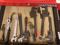 View of assorted pipe wrenches and crescent wrenches laid flat in a drawer on a woven mat with Craftsman toolbox edge visible