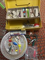 Image of open yellow Plano tackle box with compartments holding various lures, floats, and hooks as well as loose items below the box including soft plastic lures and lead weights.