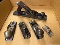 Photo of five metal wood planes in a cardboard box showing various sizes and handle types including wooden knobs and metal components