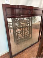Full view of the large square mirror with dark wood carved frame leaning against white wall. The frame has a carved Asian motif on the top panel. Mirror glass shows age-related spotting or patina.