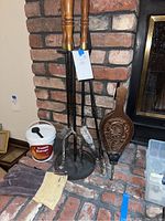 Photo showing fireplace tools with wooden handles on stand, the wooden bellows, creosote destroyer container, and gloves placed on a brick hearth near a fireplace.