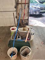 Garden tools including shovels, rakes, cultivator, broom arranged near garage door, with buckets containing small tools and accessories in front