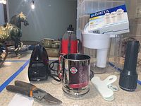 Photo showing a Brita water pitcher with installed filter cartridge, handheld electric mixer (missing beaters), Fiskars pruning shears, stainless steel flour sifter, black flashlight, white plastic bag clip, insulated water bottle, and various ceramic kitchenware near a decorative horse statue.