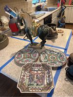 Photo showing two small round floral patterned plates, one square plate with Asian scene, and ceramic horse figurine on countertop.