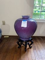 Front view of large purple porcelain vase on the black carved wooden stand on hardwood floor near window