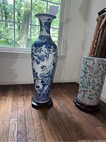 Tall blue and white oriental porcelain vase decorated with intricate floral and bird designs, placed on wooden stand by window with visible floor and background objects.