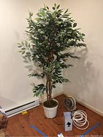 Full view showing the faux tree standing about 6 feet tall in a white plastic pot next to the small fan, spot light, and coiled cable wire on wooden floor.