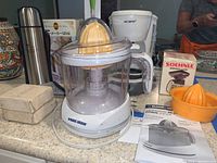 Black & Decker electric citrus juicer with clear bowl, orange juicing cone, and base on countertop with manuals and other kitchen items behind.