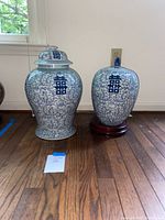 Side-by-side view of two blue and white porcelain baluster urns, one on wooden stand and both with lids.