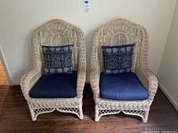 Two natural wicker chairs with navy blue cushions and Asian-inspired patterned pillows against a white wall on hardwood floor.