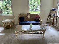 Wide shot of the wicker love seat with blue cushion and pillows centered between the wicker coffee table with glass top in front and the wicker end table with glass top to the left on beige carpet near windows.
