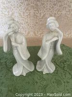 Front view of two white porcelain geisha figurines on green cloth, both standing, showing frontal pose and detail.