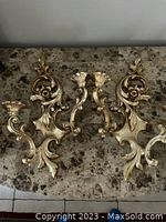 Two ornate gold finish metal wall sconces with scroll and floral motifs placed on a marble surface.