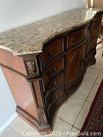 Front angle view showing granite top, ornate carved wood details, multiple drawers with knobs, and curved serpentine shape