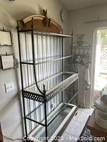 Full view of large black iron bakers rack with three glass shelves against a wall beside a window