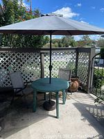 Outdoor green plastic round table with central umbrella, black weighted base, and two matching folding chairs on a patio.