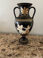 Full frontal image of black ceramic vase with two handles and Greek key and floral motifs. Central painted scene depicts Zeus on chariot with horses.