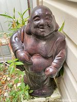 Front view of the brown ceramic Laughing Buddha statue showing the full figurine and its outdoor placement among plants.