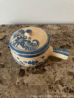 Closed view of the round clay pot with lid, showing blue floral patterns on beige background and handle.