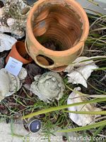 Shows large terracotta pot with round holes at top center with smaller terracotta pot and several concrete or stone outdoor ornaments around it on the ground amidst grass and wood chips.