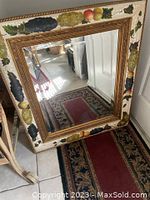 Full view of the vintage square mirror showing the ornate gold inner frame and outer cream fruit-decorated border resting on a floor with Persian-style rugs reflected