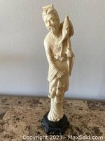 Front view of vintage carved Asian fisherman man figurine holding a fish on his shoulder, standing on a wooden pedestal.