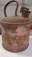 Front and top side view of antique metal gas can showing heavily rusted surface, curved handle, and spout with cap. Visible dents and faded original red paint.