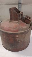 Full view of the antique metal gas can showing its rounded body and handle on top with visible patina, dents, and rust.