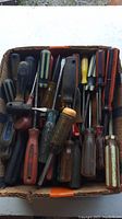 Top view showing a cardboard box filled with 40+ used screwdrivers with assorted handle colors and types, some plastic, some rubberized.