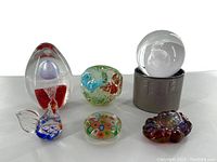 Overview of the seven paperweights on white background including egg, apple, globe, millefiori, rooster, flat iridescent, and small multicolor pieces.