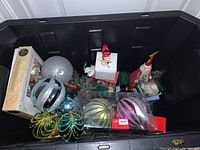 Photo of assorted Christmas ornaments and decorations in a black bin showing large colorful baubles, small figurines including Santa and dog-themed ornaments.