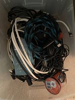 Photo of a box filled with assorted A/V cords, cables, a black electronic device, and a spool of tape.