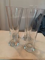 Photo showing four tall, clear, tapered beer glasses with thick bottoms arranged on a flat surface with a neutral background.