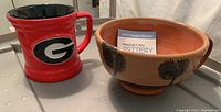Photo showing red ceramic coffee mug with University of Georgia 'G' logo and earthy colored handcrafted pottery mug with tree designs.