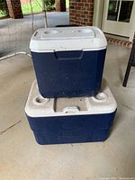Two navy blue Coleman coolers stacked, larger on bottom with wheels, smaller handheld on top, white lids with cup holders