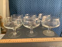Photo showing six clear etched glass champagne coupes arranged in two rows against a blue backdrop with a wooden ruler to indicate size.