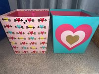 Two decorative square cardboard holiday buckets with heart motifs, one with arrow pattern and one with layered hearts